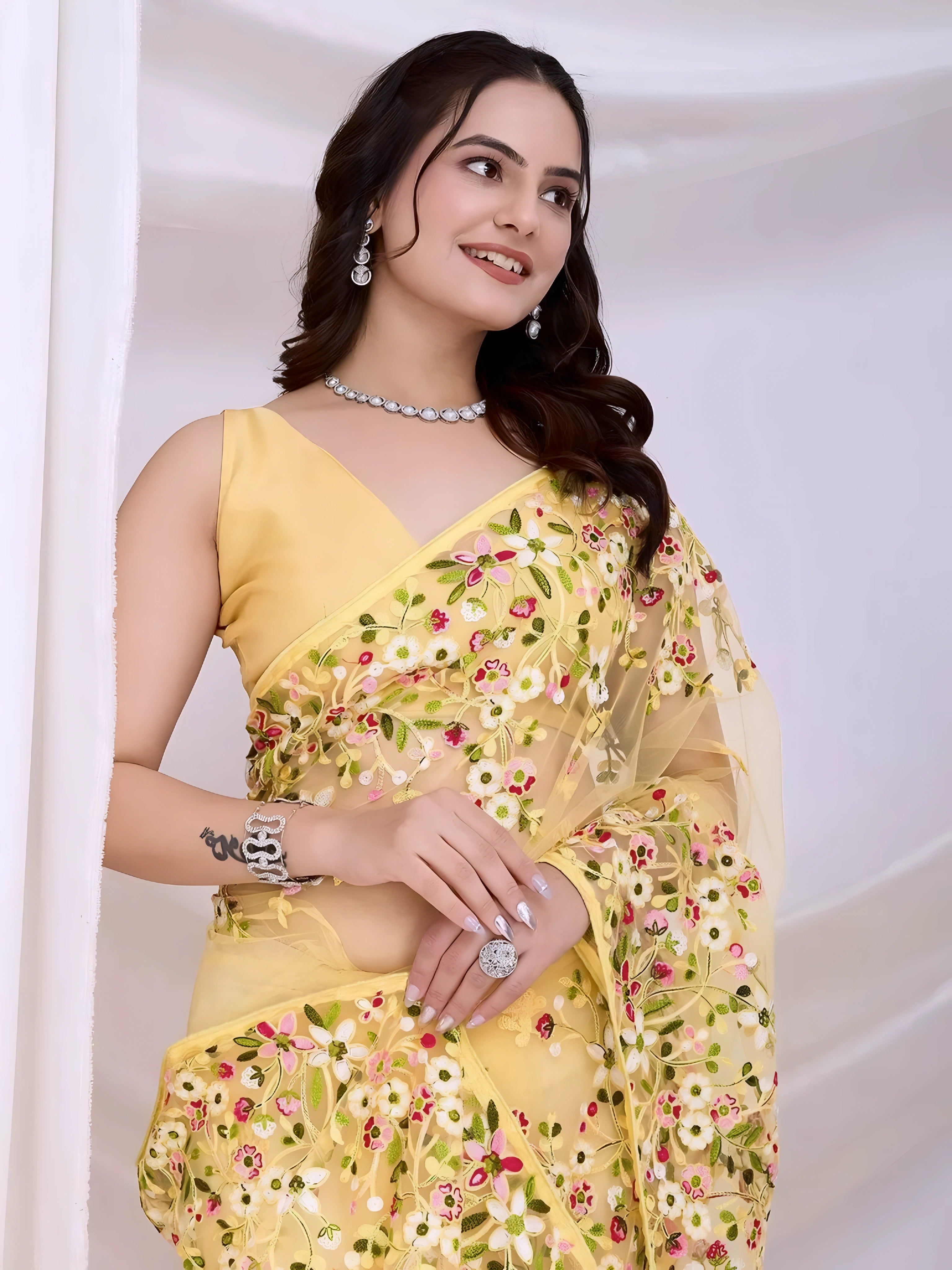 A woman models an ethereal Pastel Yellow Soft Net Saree. The sheer fabric is densely covered in delicate all-over floral embroidery featuring white, pink, and green threadwork. She wears a matching sleeveless yellow blouse and accessorizes with delicate silver/diamond jewelry, posing indoors against sheer white curtains.