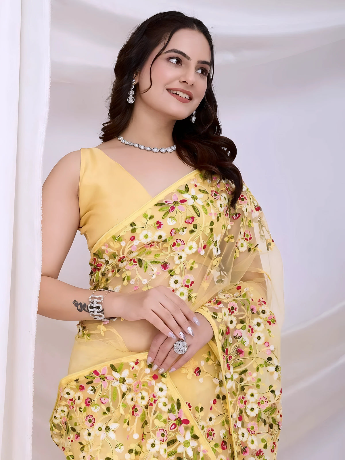 A woman models an ethereal Pastel Yellow Soft Net Saree. The sheer fabric is densely covered in delicate all-over floral embroidery featuring white, pink, and green threadwork. She wears a matching sleeveless yellow blouse and accessorizes with delicate silver/diamond jewelry, posing indoors against sheer white curtains.