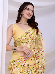A woman models an ethereal Pastel Yellow Soft Net Saree. The sheer fabric is densely covered in delicate all-over floral embroidery featuring white, pink, and green threadwork. She wears a matching sleeveless yellow blouse and accessorizes with delicate silver/diamond jewelry, posing indoors against sheer white curtains.
