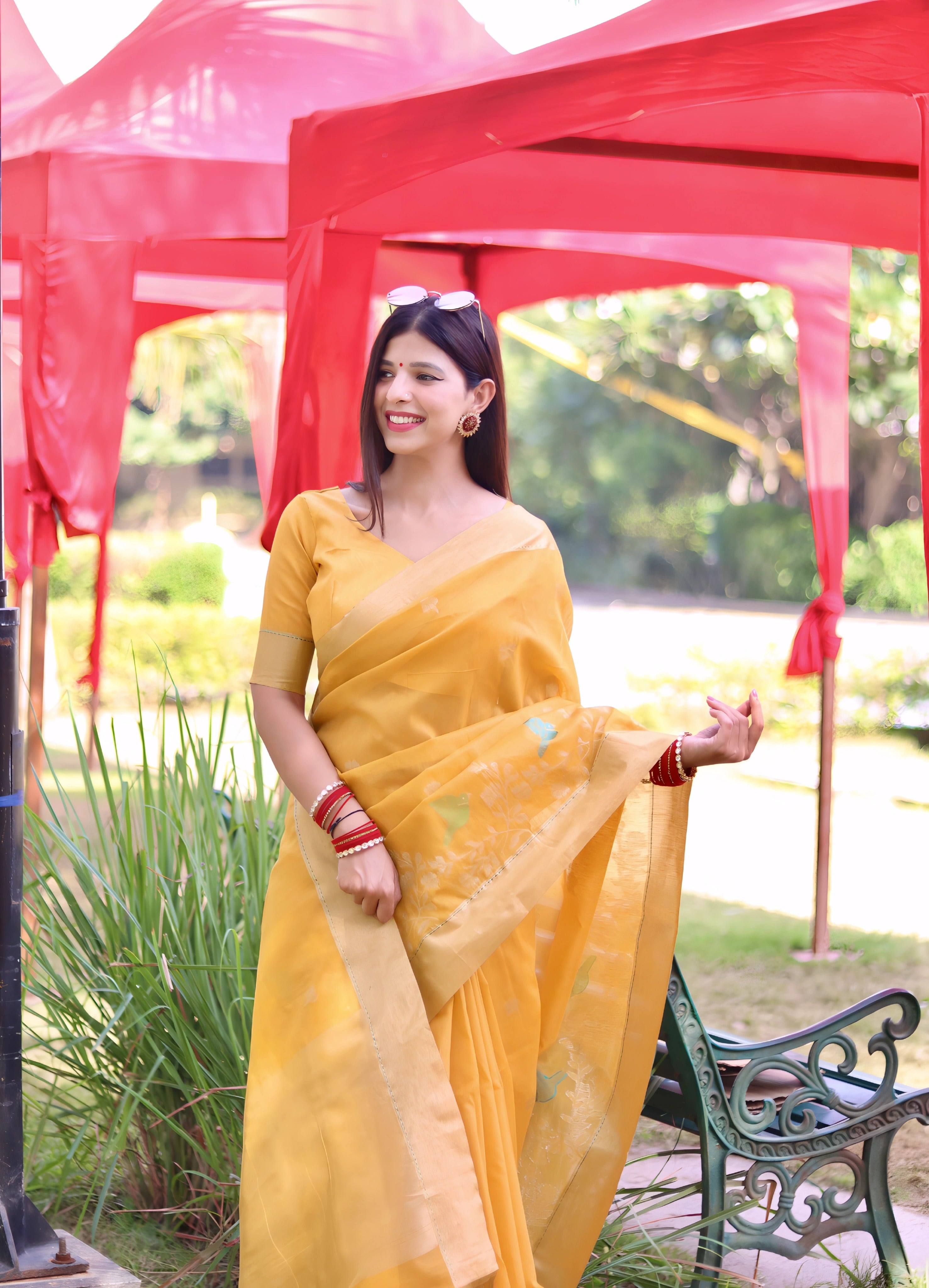 A woman models a vibrant Golden Yellow Linen Silk Saree with delicate Zari woven bird and floral motifs in mint green and blue. The saree has a wide metallic gold border. She wears striking red and orange bangles, posing outdoors under a red canopy.