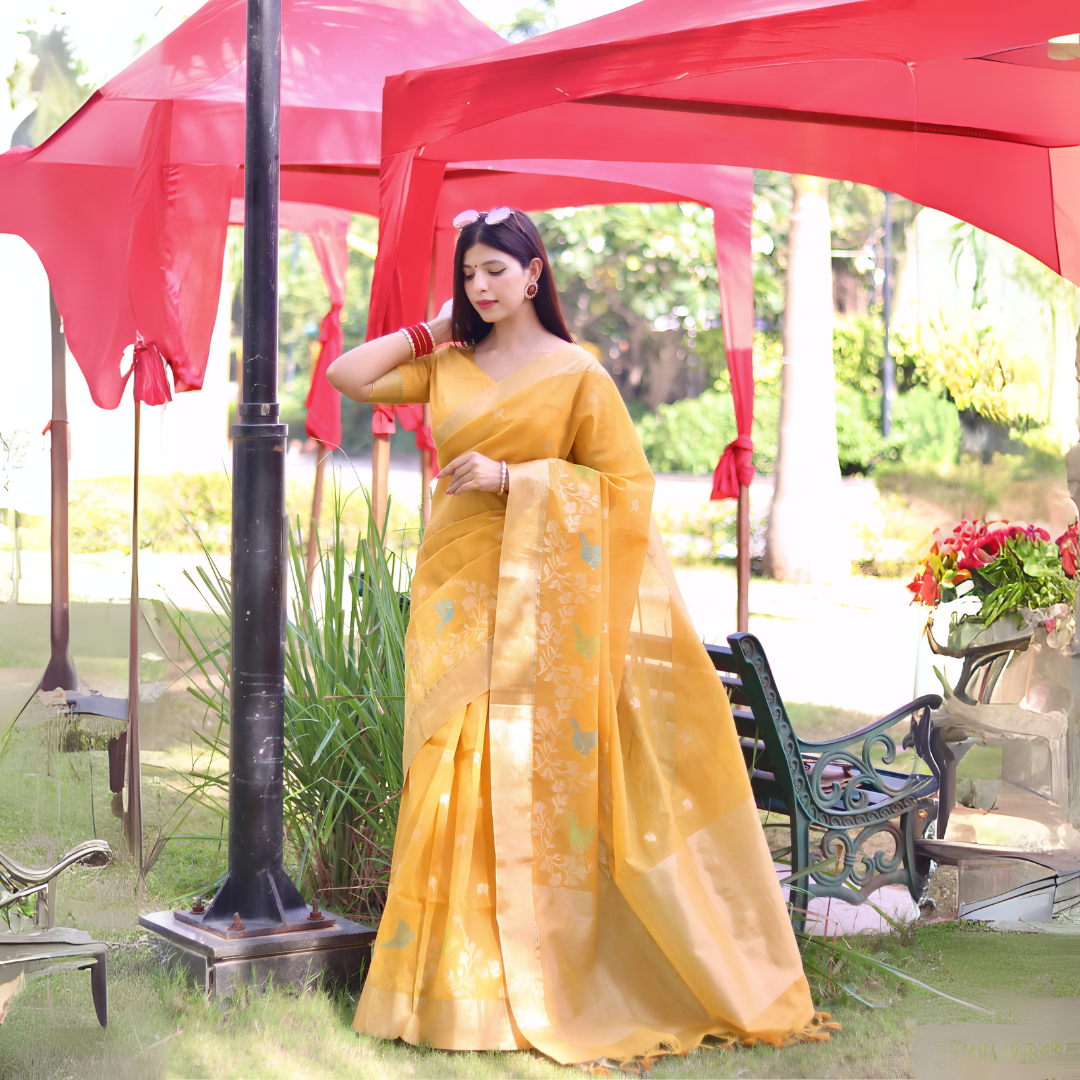 A woman models a vibrant Golden Yellow Linen Silk Saree with delicate Zari woven bird and floral motifs in mint green and blue. The saree has a wide metallic gold border. She wears striking red and orange bangles, posing outdoors under a red canopy.