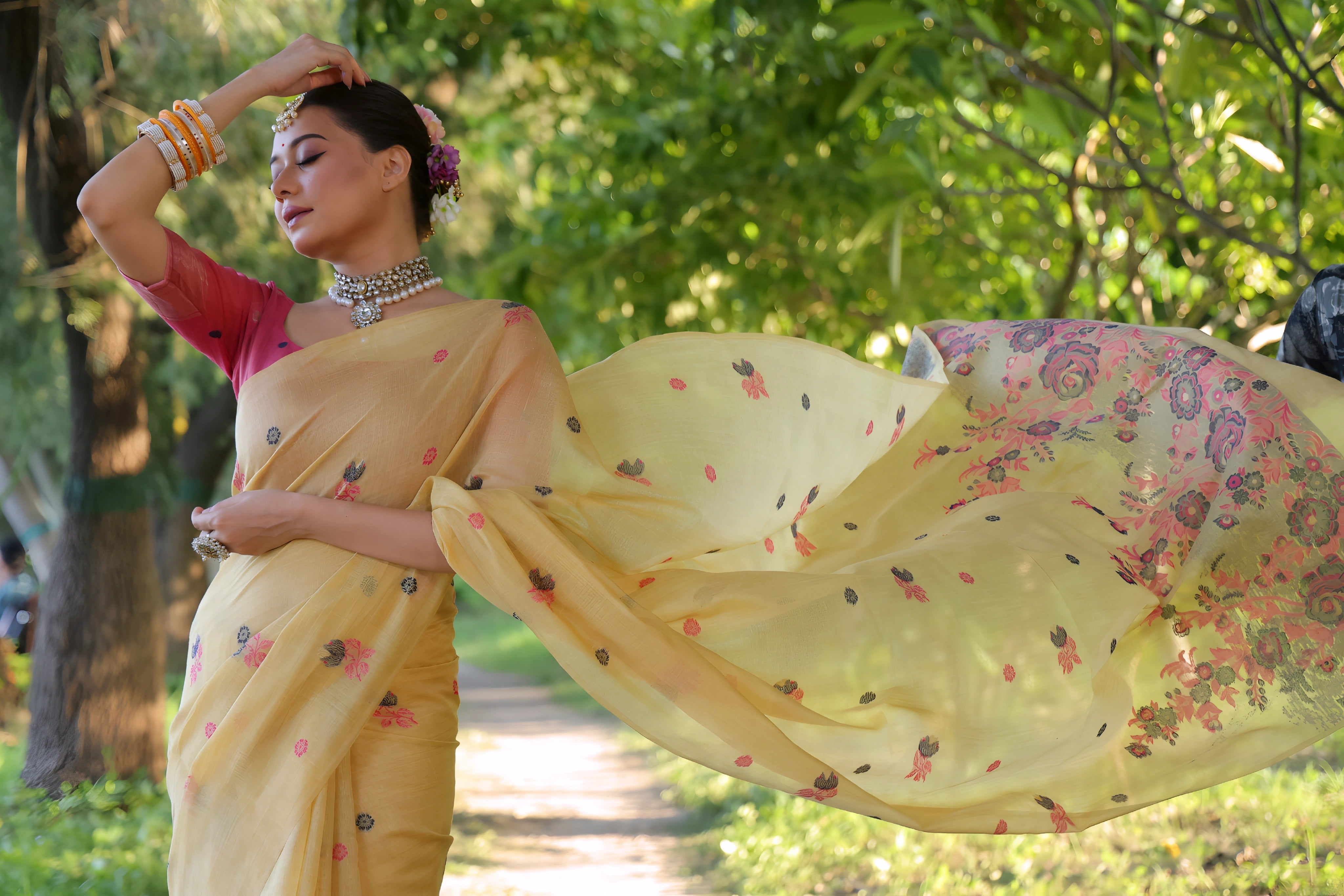A woman models an elegant Soft Pink Silk Saree with subtle texture. The saree has a wide, shimmering gold Zari border framed by deep pink piping. The pallu is the focal point, featuring rich, artistic Paithani-style print or weaving of pink, red, and brown bird-on-branch motifs. She wears a pink sleeveless blouse and gold Kundan jewelry, posing in a white, ornate courtyard.