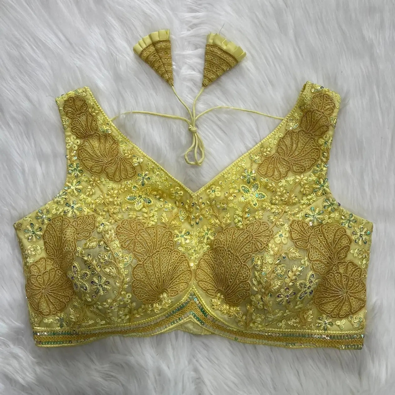 Yellow blouse featuring intricate floral and geometric sequin embroidery with gold thread detailing, deep V-neckline, and tasseled tie-back detail, displayed flat on a white fur background.
