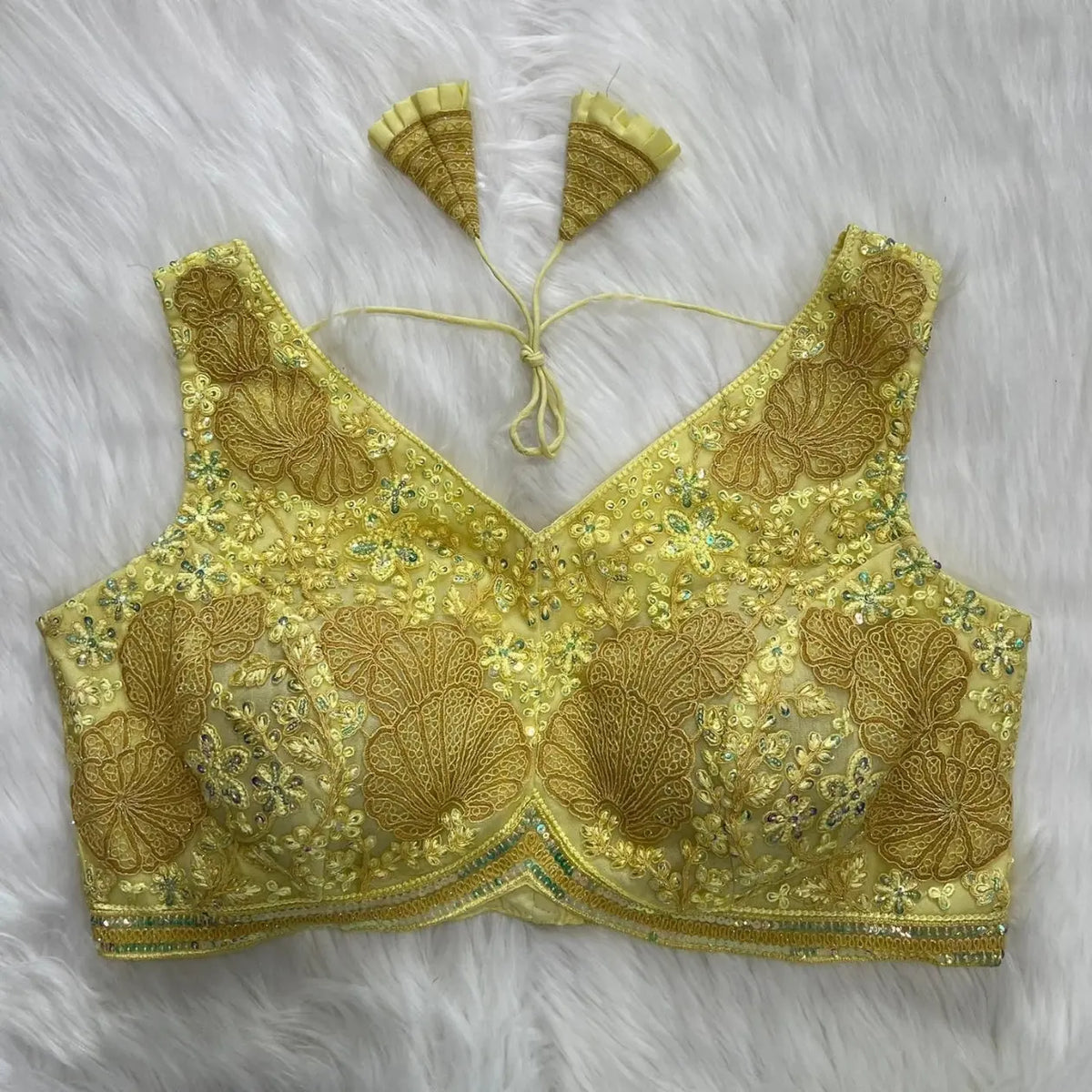 Yellow blouse featuring intricate floral and geometric sequin embroidery with gold thread detailing, deep V-neckline, and tasseled tie-back detail, displayed flat on a white fur background.
