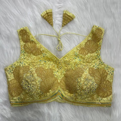 Yellow blouse featuring intricate floral and geometric sequin embroidery with gold thread detailing, deep V-neckline, and tasseled tie-back detail, displayed flat on a white fur background.