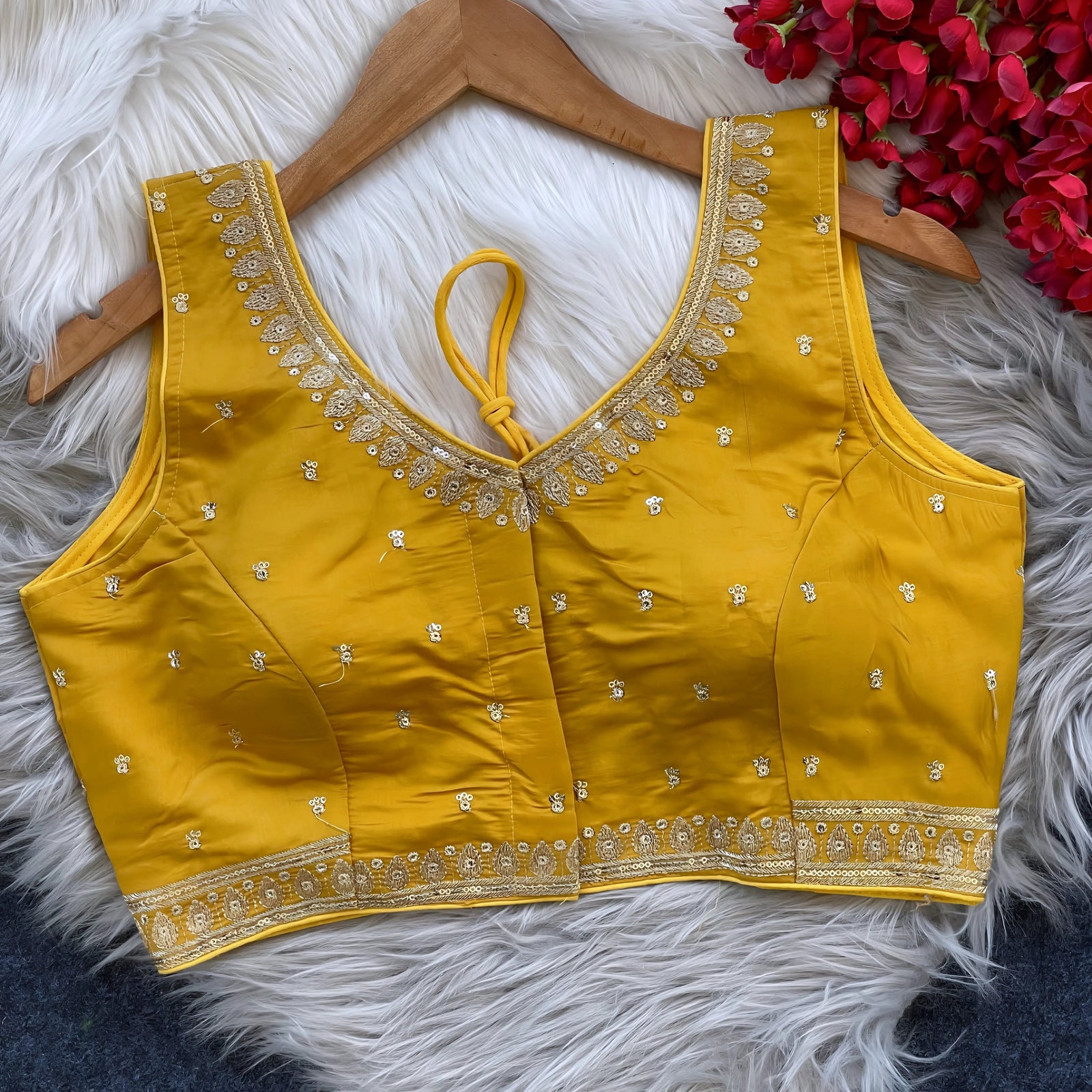 Yellow padded blouse with intricate gold embroidery along the neckline and hem, featuring small gold floral motifs, displayed on a wooden hanger over a white fur surface with red artificial flowers in the background.