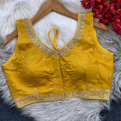 Yellow padded blouse with intricate gold embroidery along the neckline and hem, featuring small gold floral motifs, displayed on a wooden hanger over a white fur surface with red artificial flowers in the background.