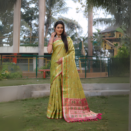 Woman wearing a yellow-green Banarasi silk saree with gold zari motifs and a vibrant pink contrast pallu featuring intricate temple and floral patterns, paired with a matching short sleeve blouse and traditional gold jewelry. She stands elegantly on a grassy lawn bordered by garden landscaping, with a background of fencing, lush trees, and playful outdoor structures, creating a graceful outdoor setting.