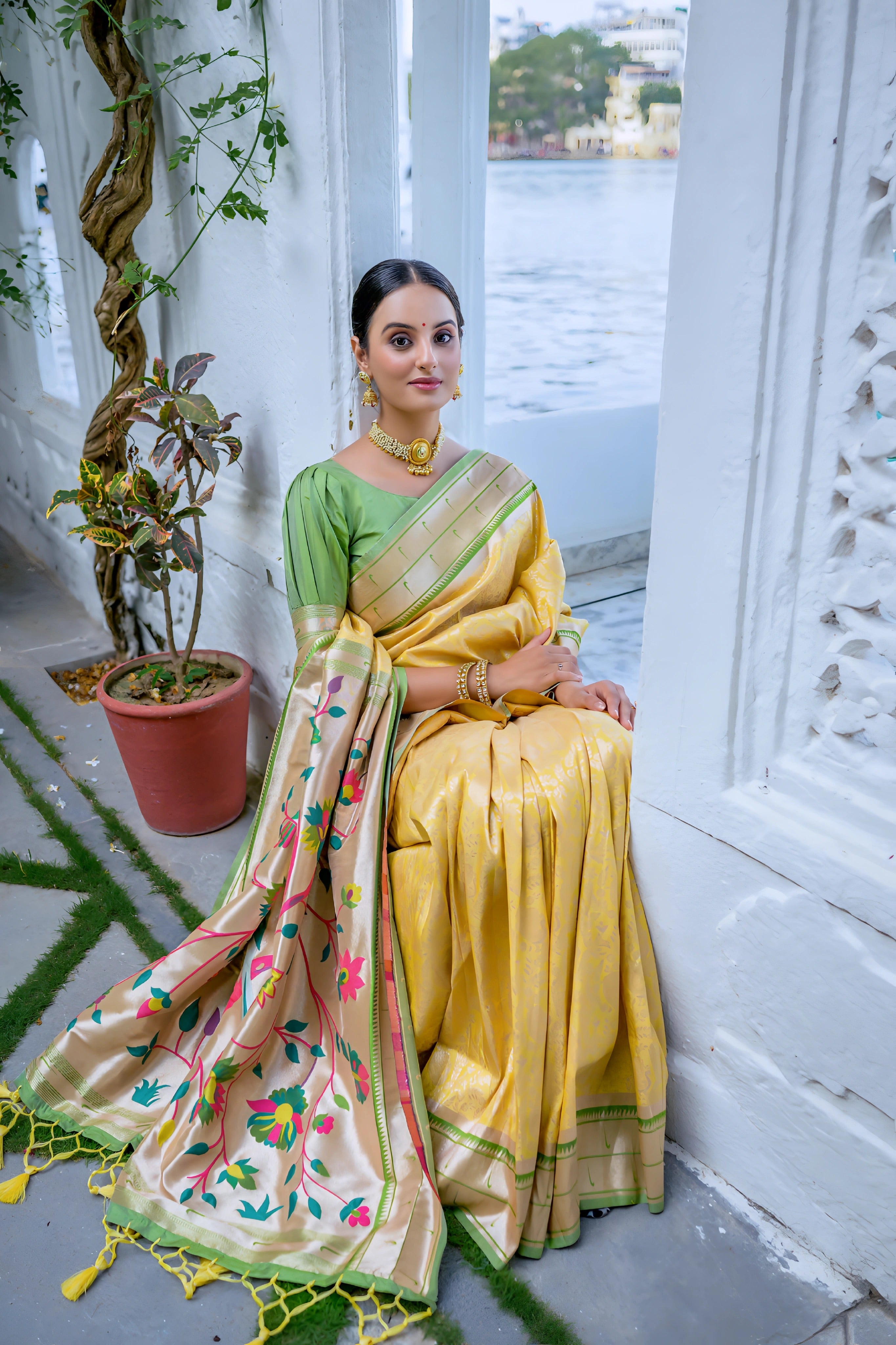 A woman models a vibrant Golden Yellow Soft Silk Saree. The saree is highlighted by a rich, wide contrast border and an elaborate pallu featuring dense, traditional Paithani Zari weaving, often depicting motifs like peacocks or florals. She is accessorized with traditional gold jewelry and posed indoors.