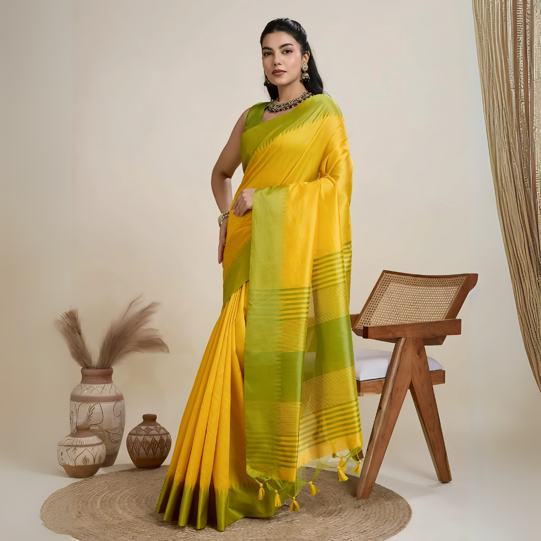 A woman models a striking Golden Yellow Raw Silk Saree. The pallu contrasts dramatically in a wide band of vibrant Lime Green with horizontal stripe weaving. She wears a sleeveless green blouse and accessorizes with a chunky dark gemstone necklace and large earrings, posing indoors.