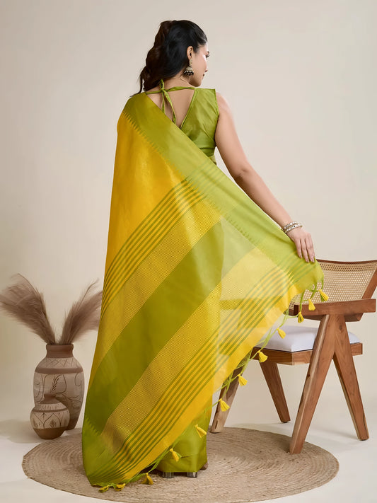 A woman models a striking Golden Yellow Raw Silk Saree. The pallu contrasts dramatically in a wide band of vibrant Lime Green with horizontal stripe weaving. She wears a sleeveless green blouse and accessorizes with a chunky dark gemstone necklace and large earrings, posing indoors.