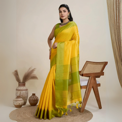 A woman models a striking Golden Yellow Raw Silk Saree. The pallu contrasts dramatically in a wide band of vibrant Lime Green with horizontal stripe weaving. She wears a sleeveless green blouse and accessorizes with a chunky dark gemstone necklace and large earrings, posing indoors.