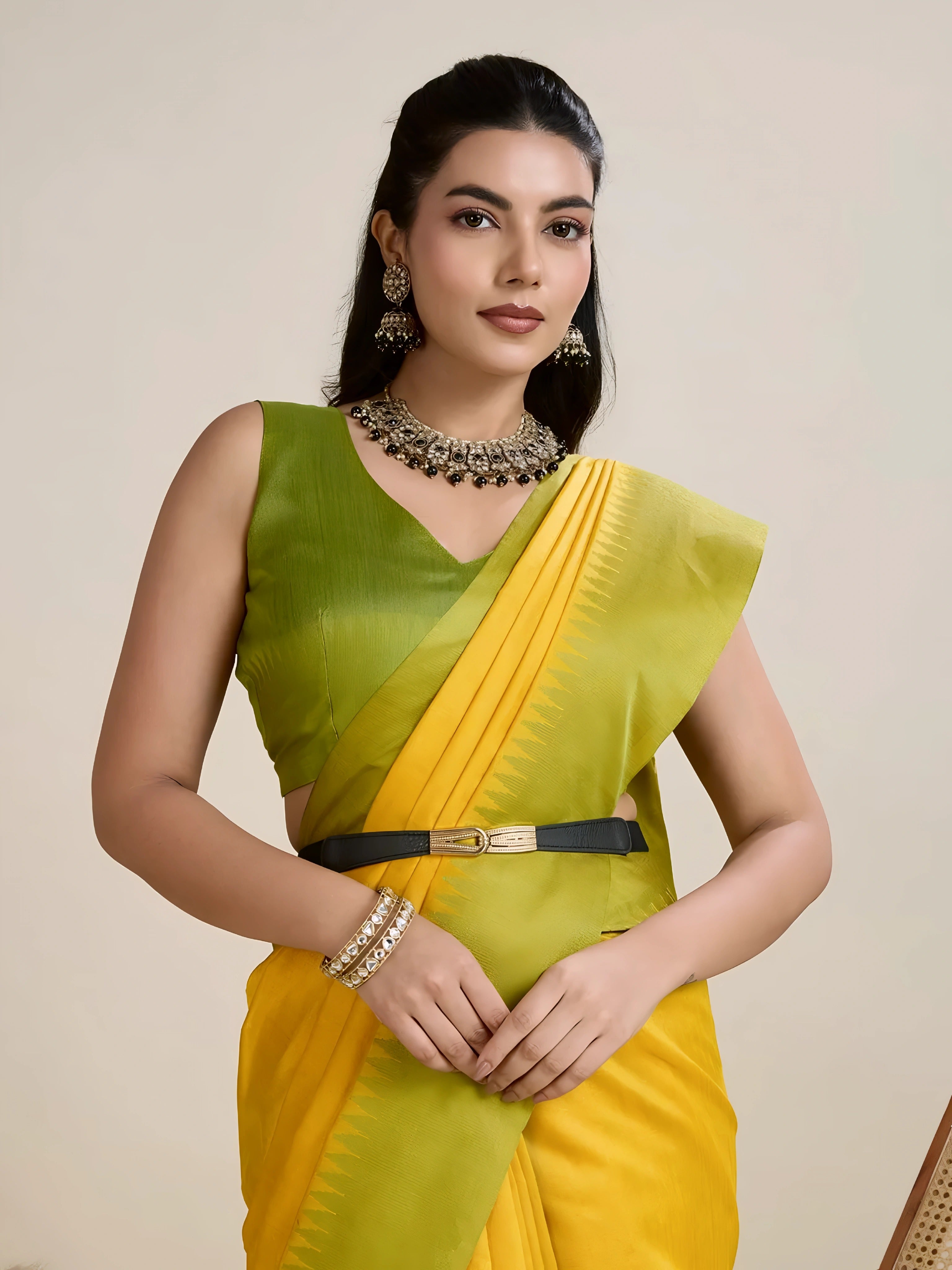 A woman models a striking Golden Yellow Raw Silk Saree. The pallu contrasts dramatically in a wide band of vibrant Lime Green with horizontal stripe weaving. She wears a sleeveless green blouse and accessorizes with a chunky dark gemstone necklace and large earrings, posing indoors.