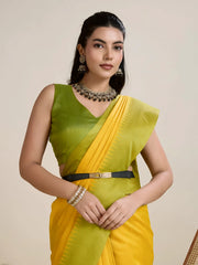 A woman models a striking Golden Yellow Raw Silk Saree. The pallu contrasts dramatically in a wide band of vibrant Lime Green with horizontal stripe weaving. She wears a sleeveless green blouse and accessorizes with a chunky dark gemstone necklace and large earrings, posing indoors.
