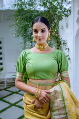 A woman models a vibrant Golden Yellow Soft Silk Saree. The saree is highlighted by a rich, wide contrast border and an elaborate pallu featuring dense, traditional Paithani Zari weaving, often depicting motifs like peacocks or florals. She is accessorized with traditional gold jewelry and posed indoors.