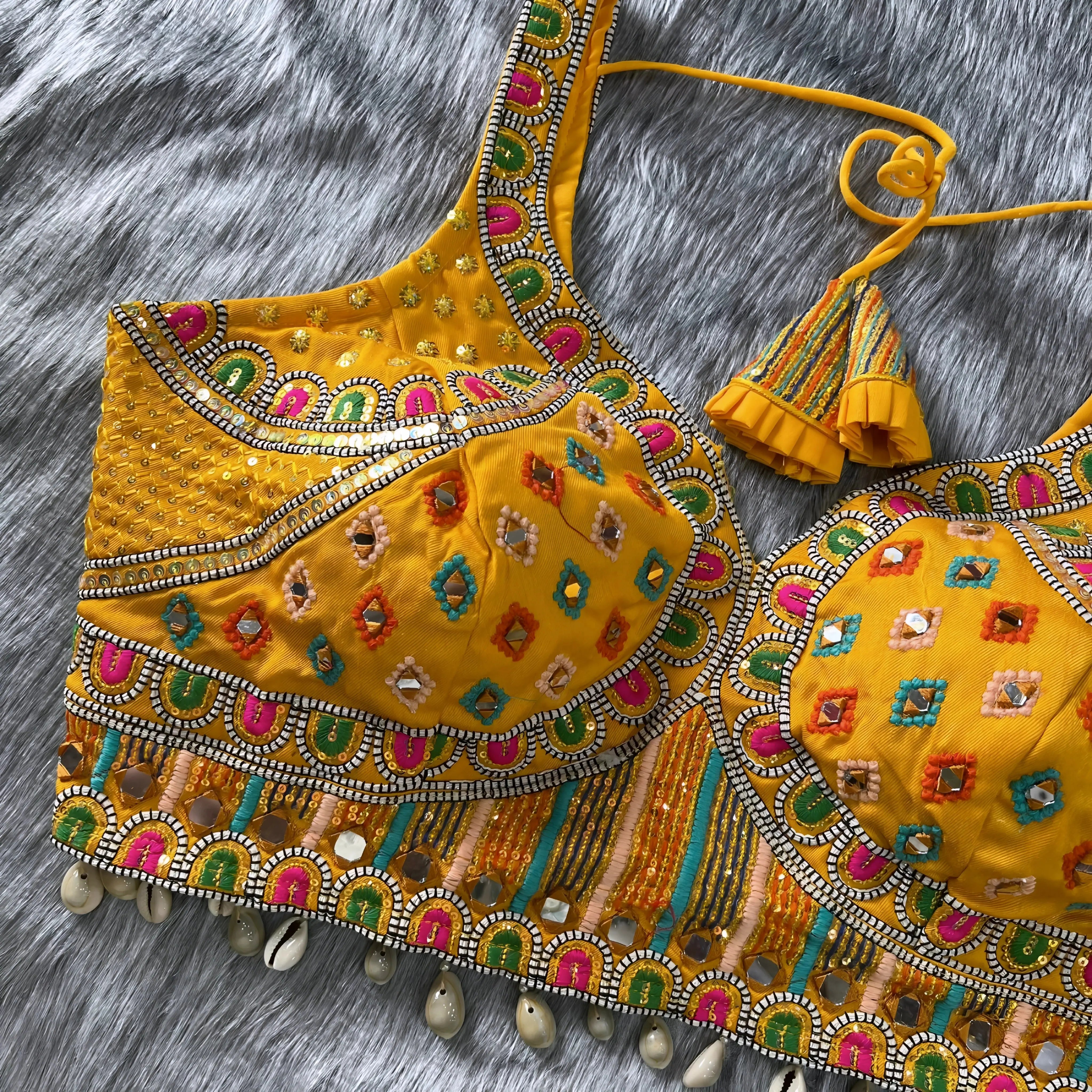 Traditional yellow Gujarati blouse featuring intricate multicolor geometric embroidery, mirror work, and bead accents, with a decorative shell trim along the hem. The structured cups, wide straps, and tasseled yellow tie-back detail are displayed flat on a soft grey fur background.