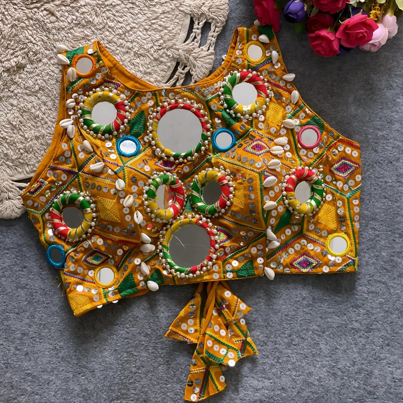 Traditional yellow Kutch-style blouse richly adorned with circular mirror work, multicolor rings, geometric thread embroidery, and decorative cowrie shells, featuring tasseled tie-back detail and displayed flat on a grey textured surface beside a braided cream mat and a bouquet of vibrant artificial roses.