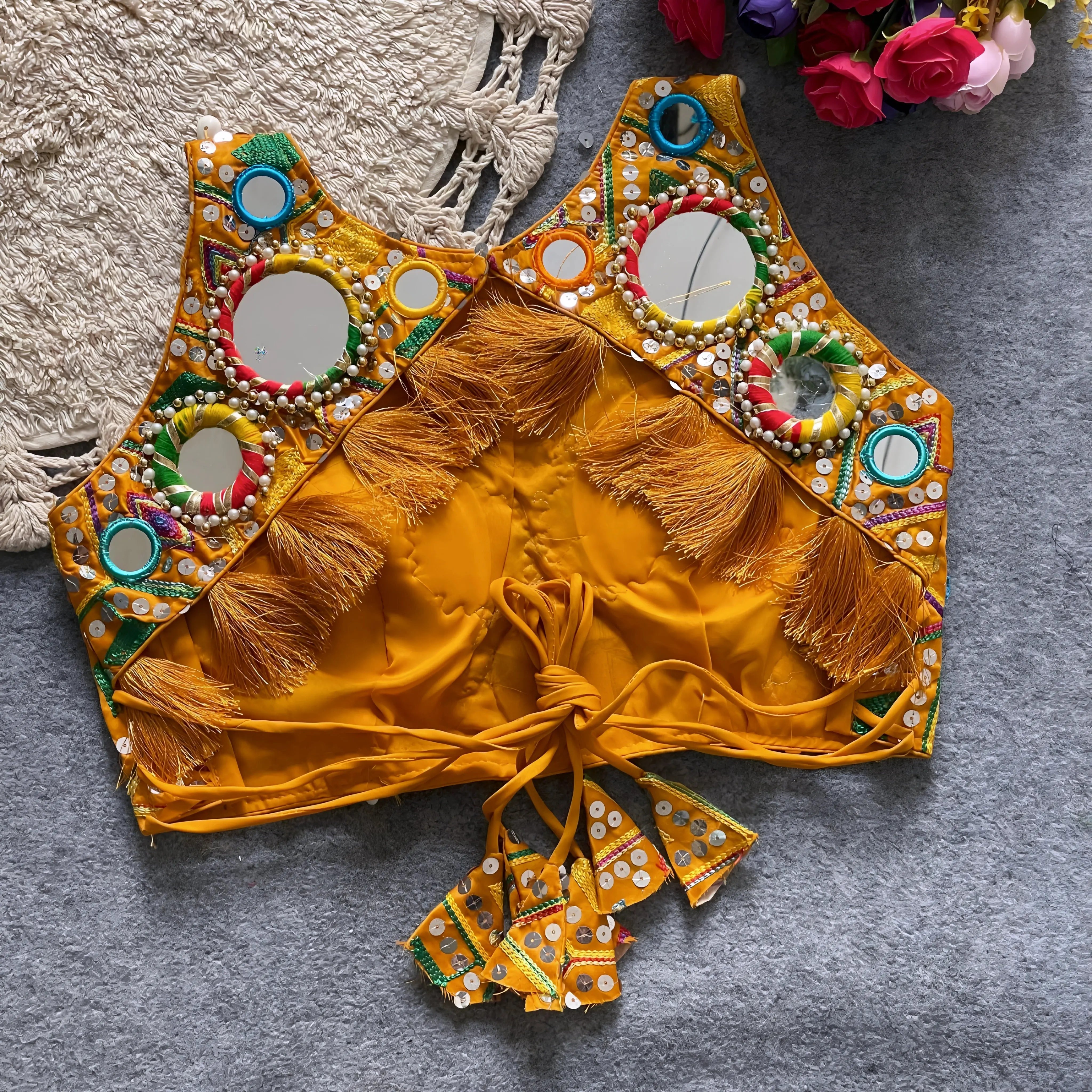 Traditional yellow Kutch-style blouse richly adorned with circular mirror work, multicolor rings, geometric thread embroidery, and decorative cowrie shells, featuring tasseled tie-back detail and displayed flat on a grey textured surface beside a braided cream mat and a bouquet of vibrant artificial roses.