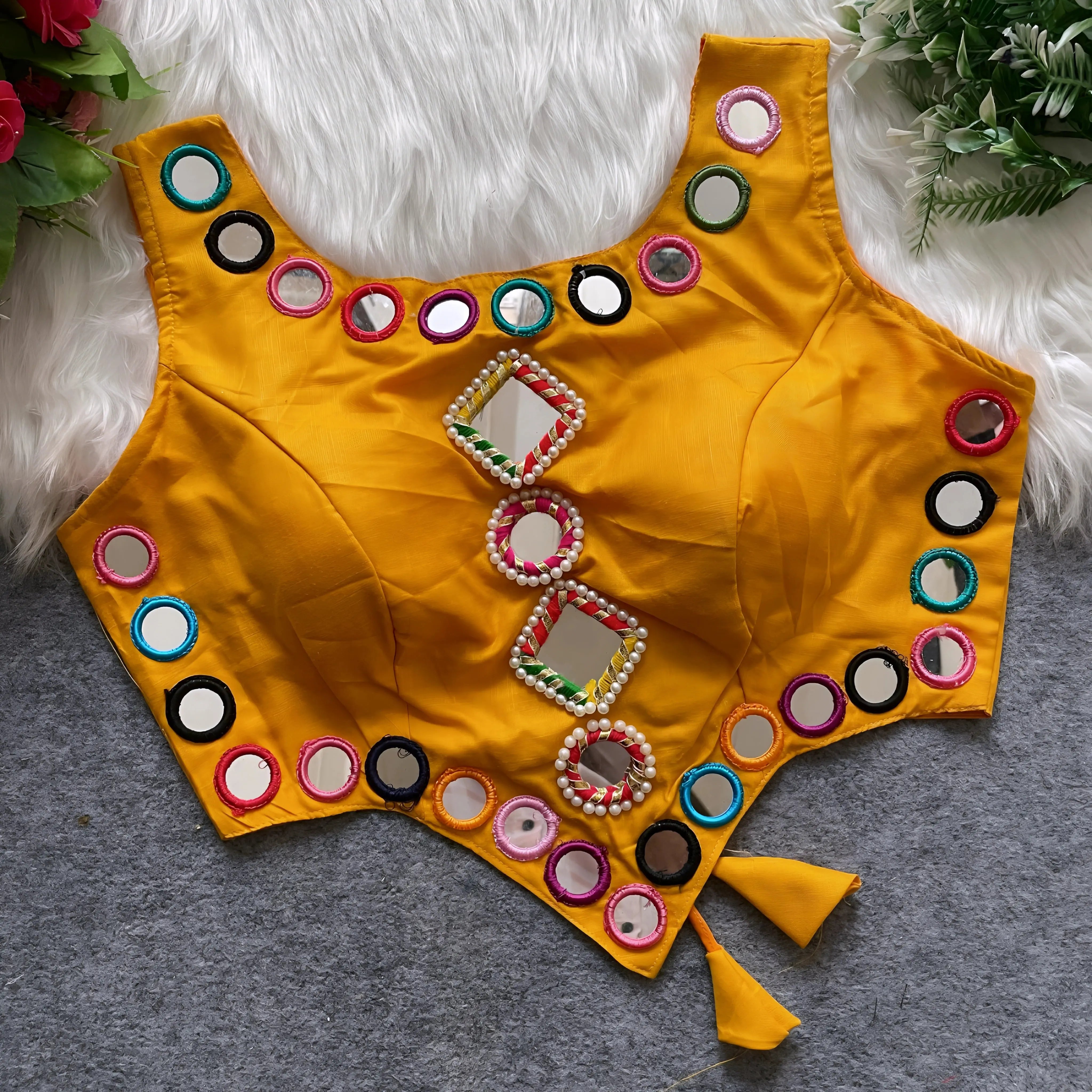 Bright yellow ethnic blouse featuring intricate mirror work with multicolored embroidered borders and diamond and round mirrors at the center, adorned with decorative pearl details and side tie-up tassels. Displayed flat on a grey surface with white faux fur and green leafy accents, highlighting its festive and traditional design.
