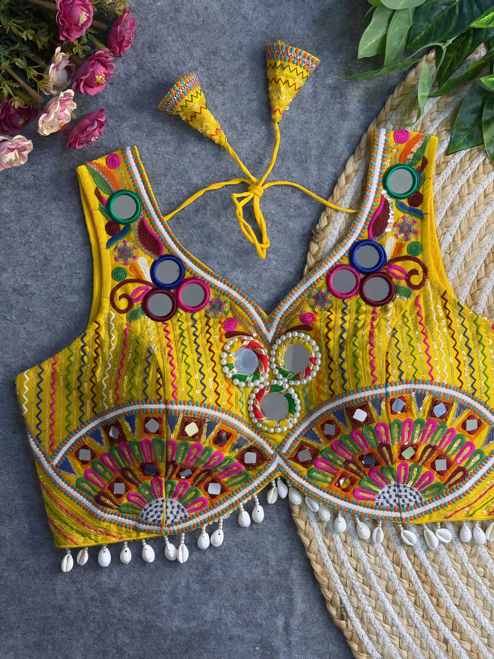 Traditional yellow embroidered blouse featuring vibrant multicolor mirror work, intricate thread embroidery in geometric and floral patterns, and shell tassel detailing along the hem, displayed on a flat surface with floral and leafy decor.