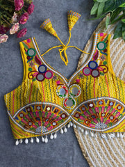 Traditional yellow embroidered blouse featuring vibrant multicolor mirror work, intricate thread embroidery in geometric and floral patterns, and shell tassel detailing along the hem, displayed on a flat surface with floral and leafy decor.