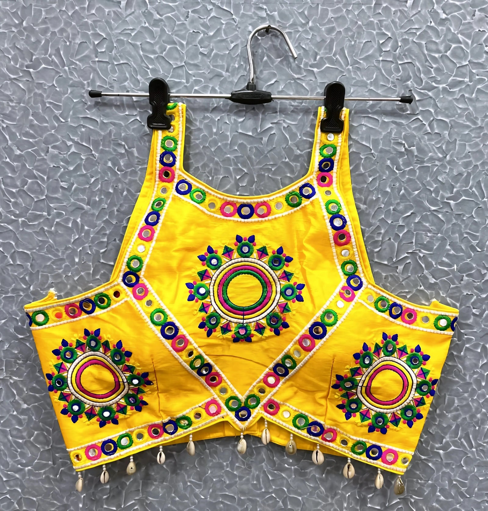 Yellow blouse featuring vibrant circular and oval mirror work with traditional Kutch multicolor embroidery, accented by geometric borders and shell tassel drops along the hem. Displayed on a hanger against a textured silver background, showcasing its festive and culturally inspired ethnic design.Yellow blouse featuring vibrant circular and oval mirror work with traditional Kutch multicolor embroidery, accented by geometric borders and shell tassel drops along the hem. Displayed on a hanger against a texture