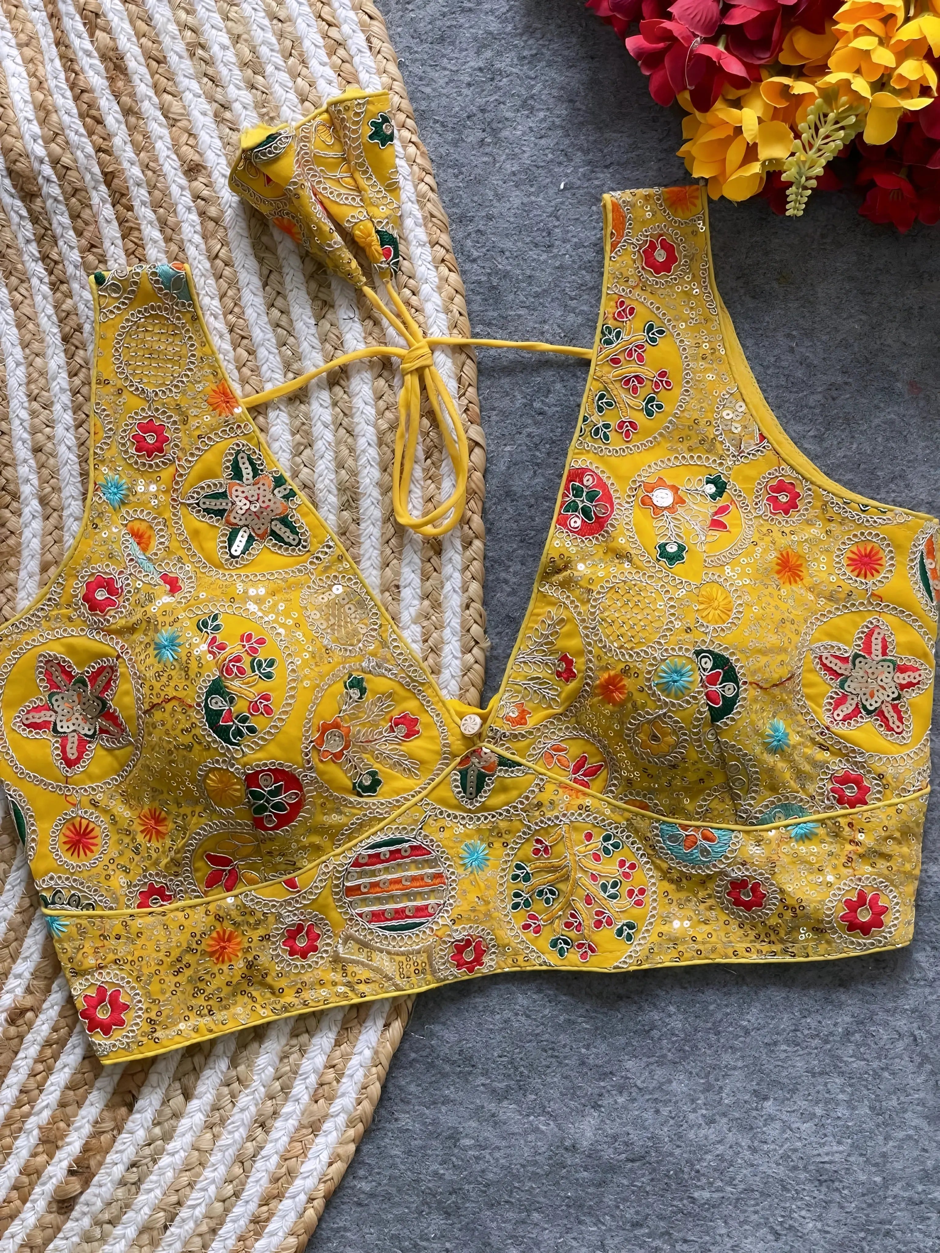 Traditional yellow blouse featuring vibrant multicolor thread embroidery and mirror work in circular floral and geometric patterns, with sequin accents and a deep V-neck tie-back detail, displayed flat on a textured mat with artificial red and yellow flowers in the corner.