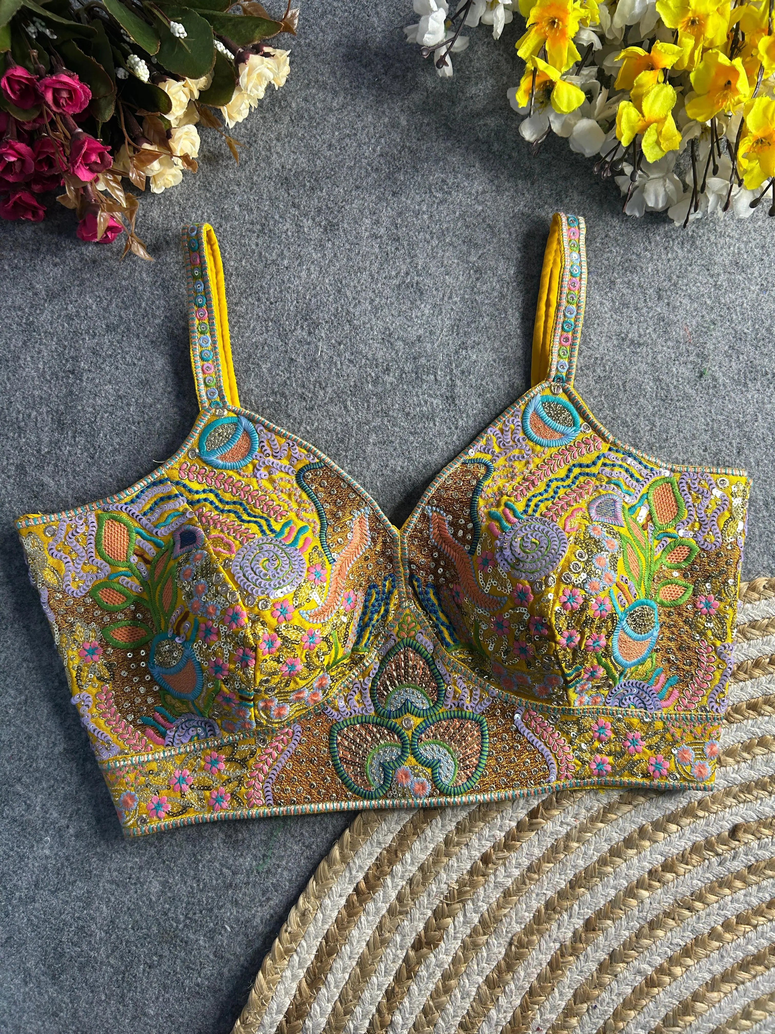 Traditional yellow bustier-style blouse featuring vibrant multicolor Madhubani-inspired embroidery with intricate floral, paisley, and geometric motifs, embellished with sequins and beads, structured cups, and adjustable straps, displayed flat on a textured grey surface with artificial pink, yellow, and white flowers in the corners.

Traditional yellow bustier-style blouse featuring vibrant multicolor Madhubani-inspired embroidery with intricate floral, paisley, and geometric motifs, embellished with sequin