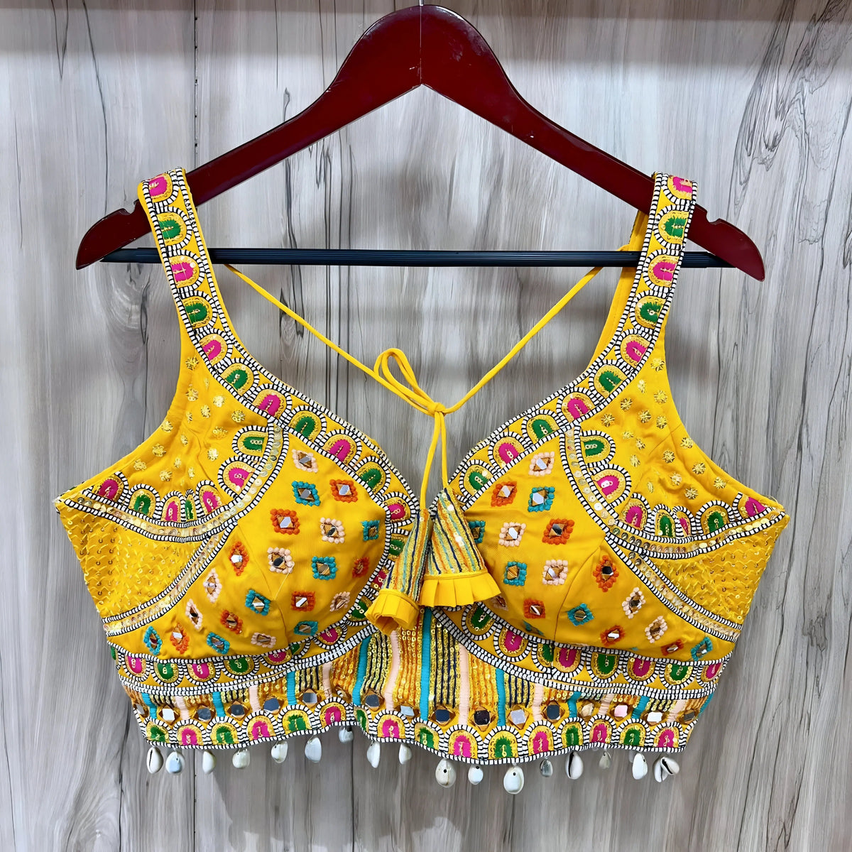Traditional yellow Kutch-style blouse beautifully adorned with vibrant multicolor geometric embroidery, detailed mirror work including circular and teardrop shapes, accented with colorful beads and cowrie shell trim at the hem. The blouse features wide straps, structured cups, and tasseled yellow tie-back details, displayed flat on a grey textured surface with artificial pink flowers and green foliage at the corners.