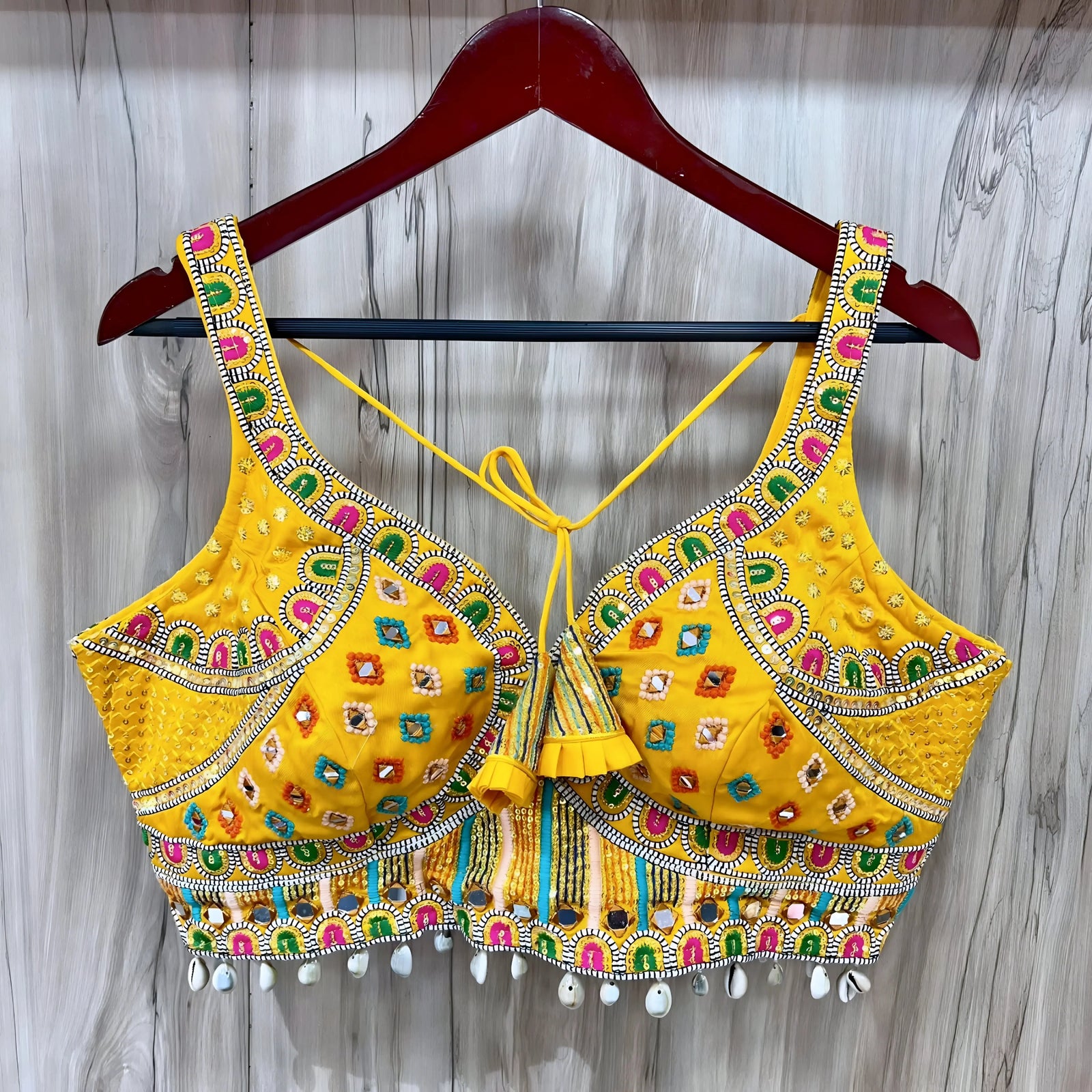 Traditional yellow Kutch-style blouse beautifully adorned with vibrant multicolor geometric embroidery, detailed mirror work including circular and teardrop shapes, accented with colorful beads and cowrie shell trim at the hem. The blouse features wide straps, structured cups, and tasseled yellow tie-back details, displayed flat on a grey textured surface with artificial pink flowers and green foliage at the corners.
