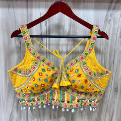 Traditional yellow Kutch-style blouse beautifully adorned with vibrant multicolor geometric embroidery, detailed mirror work including circular and teardrop shapes, accented with colorful beads and cowrie shell trim at the hem. The blouse features wide straps, structured cups, and tasseled yellow tie-back details, displayed flat on a grey textured surface with artificial pink flowers and green foliage at the corners.
