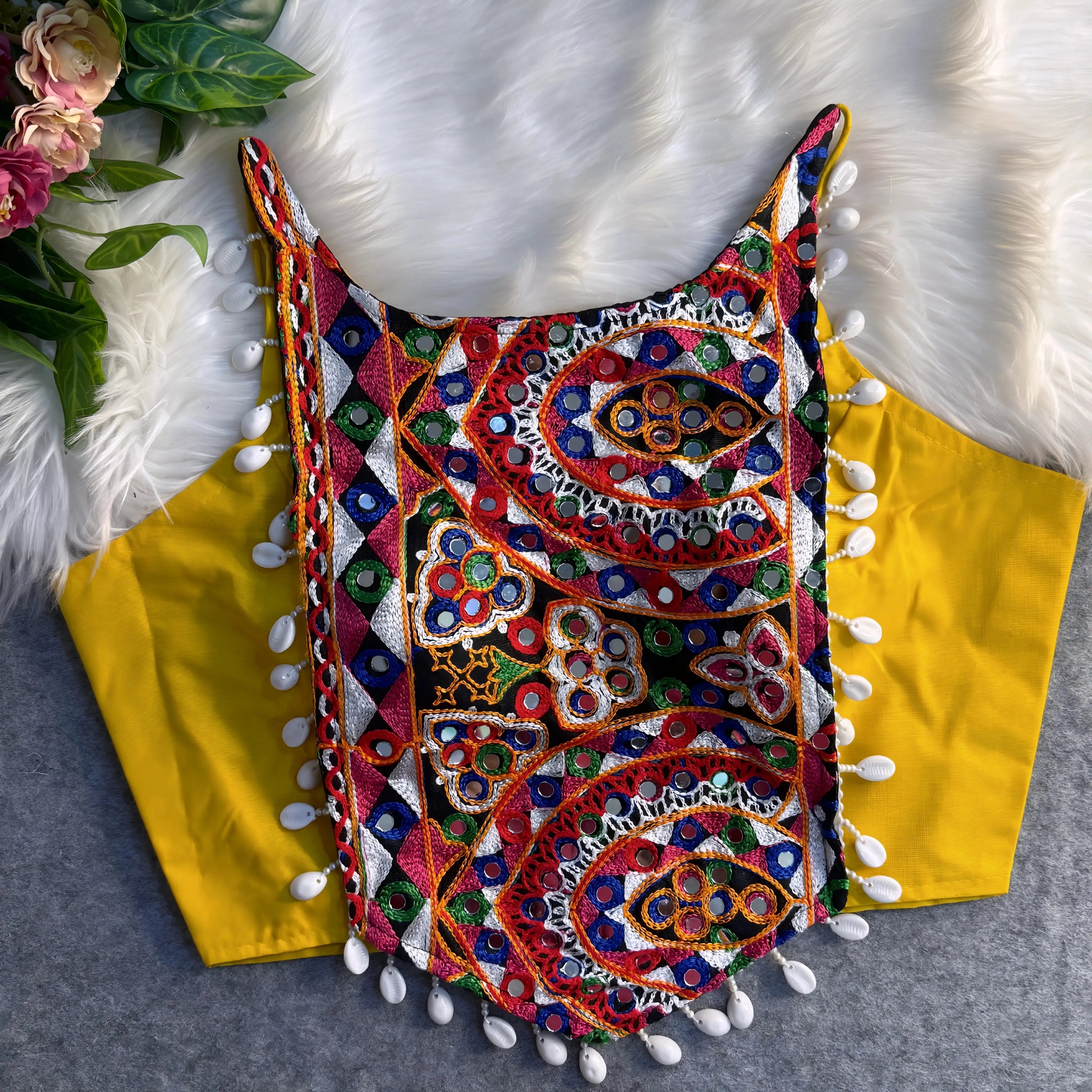 Traditional yellow blouse featuring vibrant multicolor mirror work and intricate thread embroidery in geometric and paisley patterns, accented with white shell tassels along the hem, displayed flat on a soft fur and grey background with floral and leafy decor.