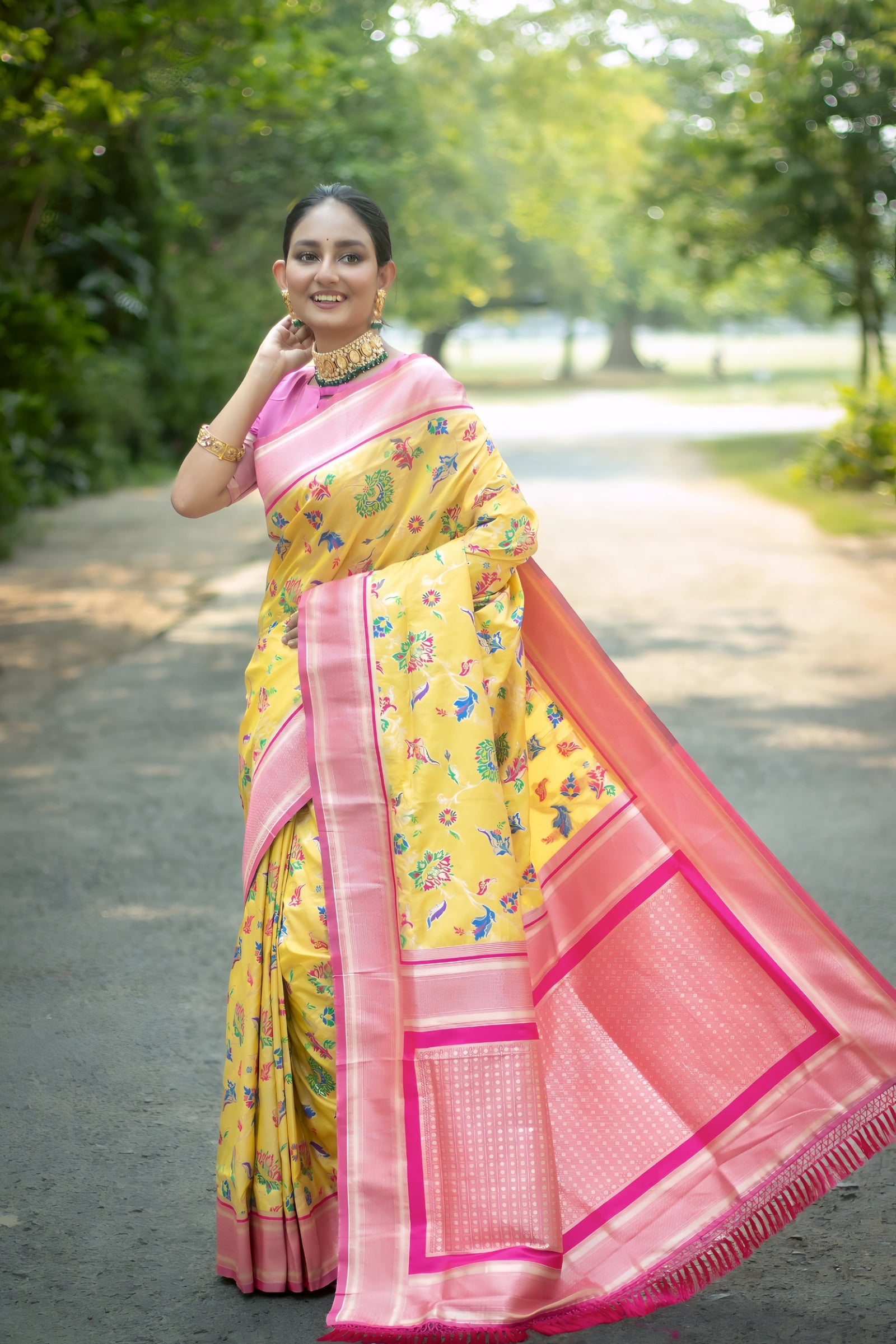 Woman wearing a yellow silk saree with pink borders and multicolor floral motifs, paired with a pink blouse and traditional jewelry, standing gracefully on a tree-lined garden path with soft natural light and lush greenery in the background.
