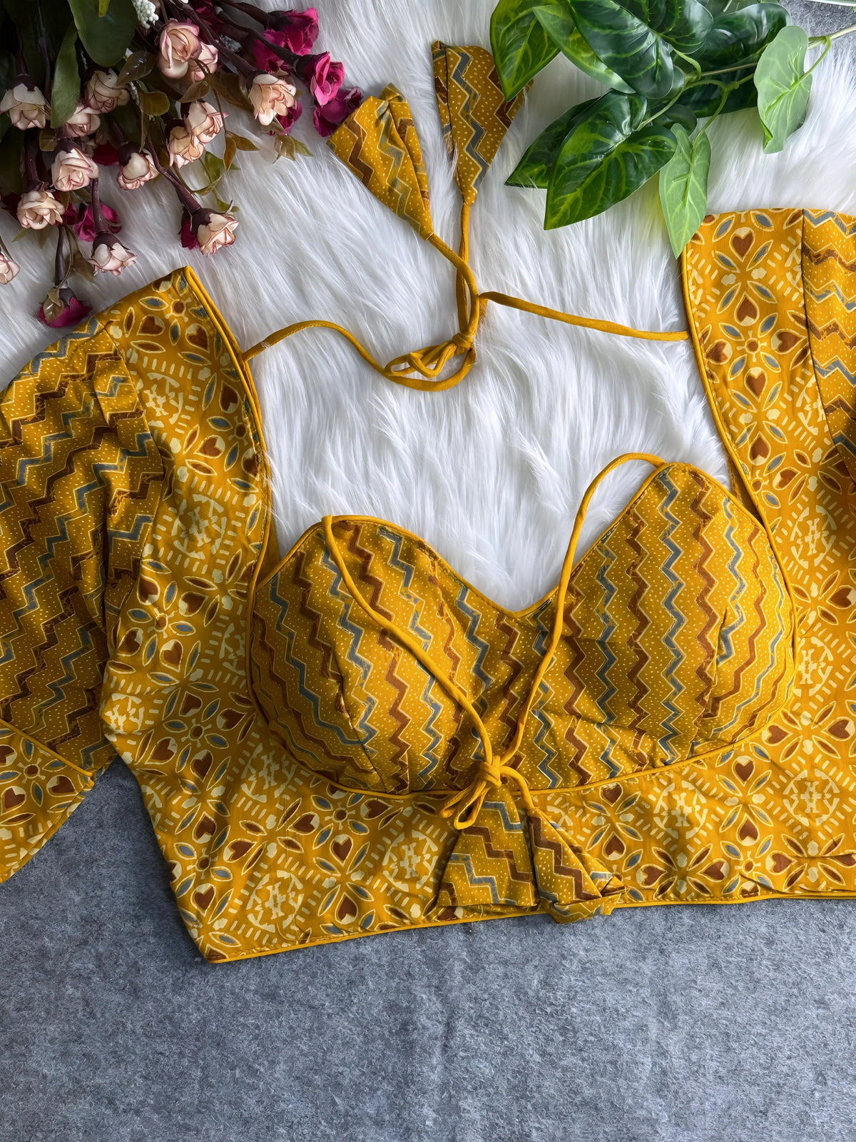 Yellow printed padded blouse with geometric and floral patterns, displayed on a white fur surface with artificial flowers and green leaves in the background.