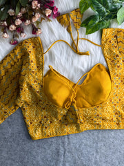 Yellow printed padded blouse with geometric and floral patterns, displayed on a white fur surface with artificial flowers and green leaves in the background.
