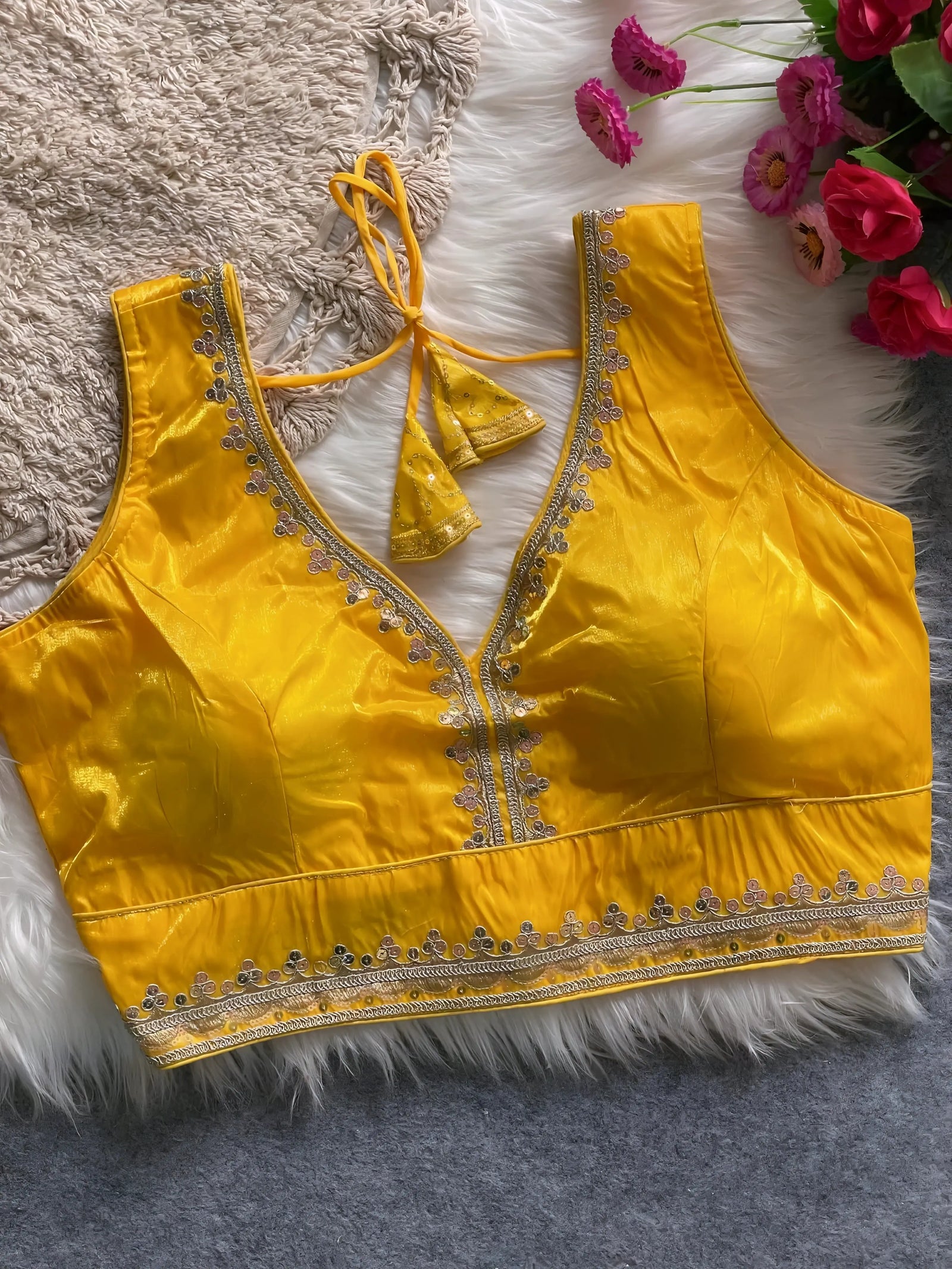 Traditional yellow silk blouse featuring intricate gold sequin and thread embroidery along the neckline and hem, with a decorative tassel tie at the back, displayed flat on a soft fur and textured mat background with artificial pink flowers in the corner.