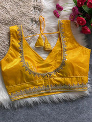 Traditional yellow silk blouse featuring intricate gold sequin and thread embroidery along the neckline and hem, with a decorative tassel tie at the back, displayed flat on a soft fur and textured mat background with artificial pink flowers in the corner.