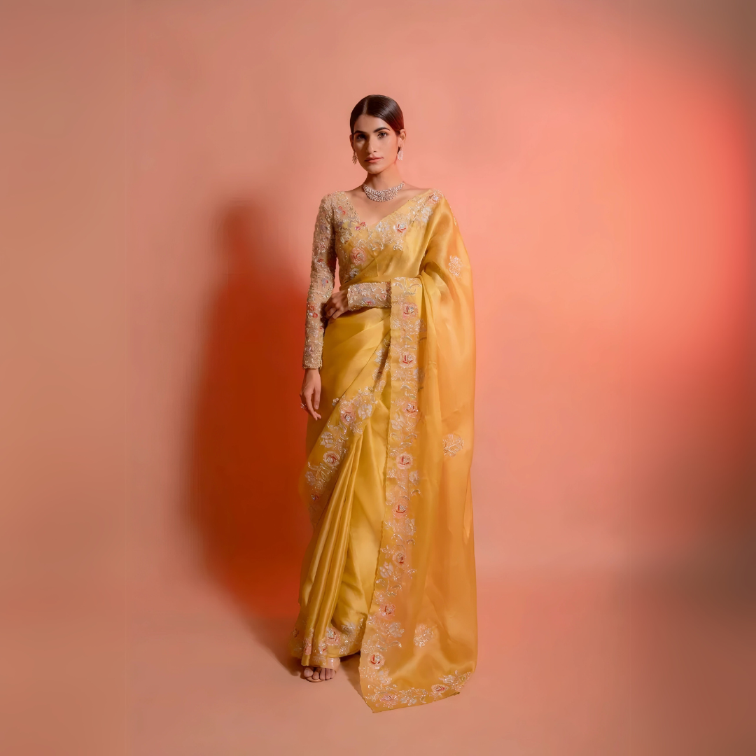 A woman models a striking Golden Yellow Soft Organza Silk Saree. The saree features delicate thread and sequin floral embroidery along the border. She wears a matching long-sleeve, heavily embroidered blouse and accessorizes with silver/diamond jewelry.