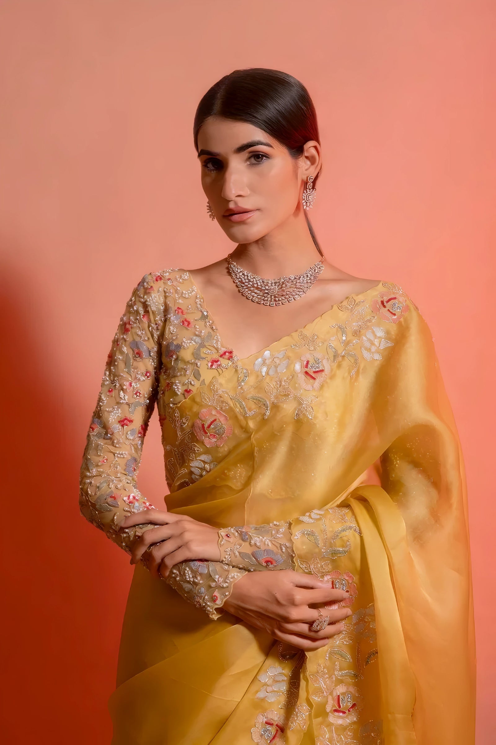 A woman models a striking Golden Yellow Soft Organza Silk Saree. The saree features delicate thread and sequin floral embroidery along the border. She wears a matching long-sleeve, heavily embroidered blouse and accessorizes with silver/diamond jewelry.