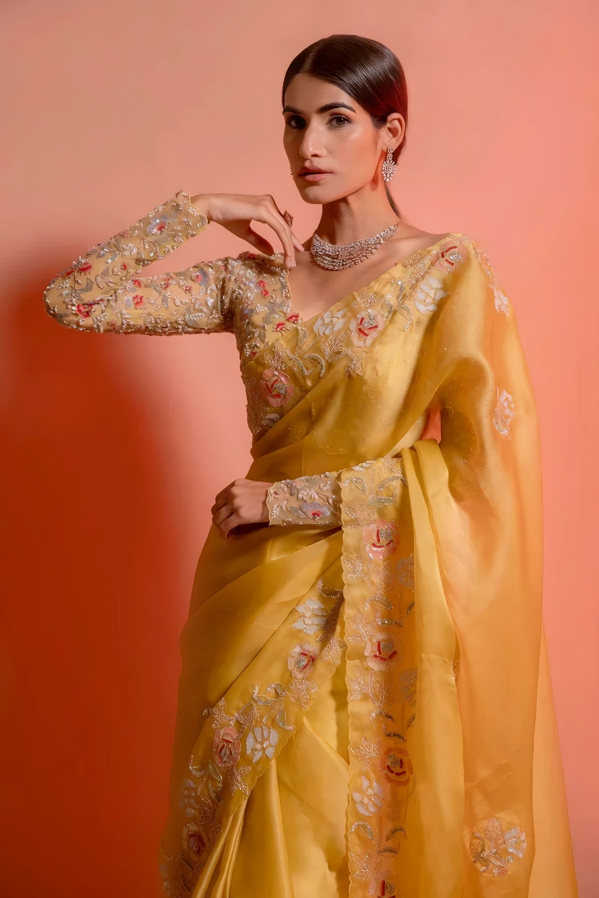 A woman models a striking Golden Yellow Soft Organza Silk Saree. The saree features delicate thread and sequin floral embroidery along the border. She wears a matching long-sleeve, heavily embroidered blouse and accessorizes with silver/diamond jewelry.