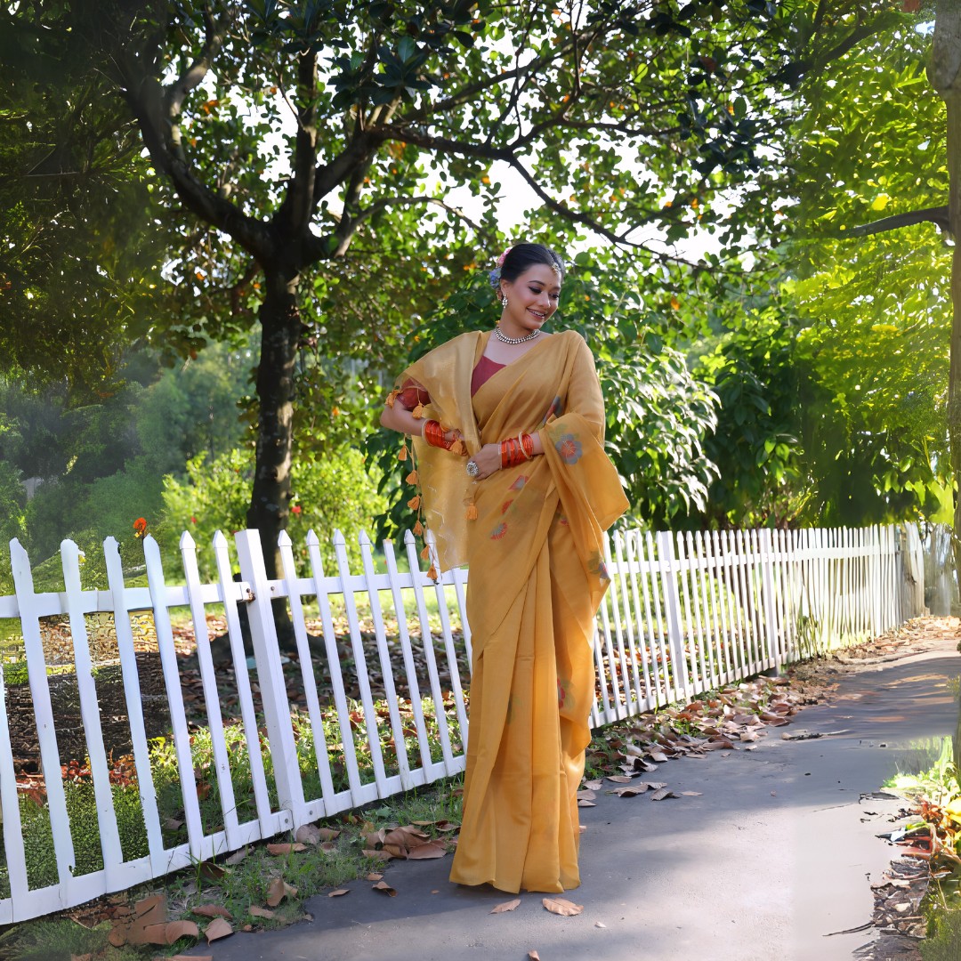 A woman models a vibrant Mustard Yellow Soft Muga Cotton Saree. The pallu features delicate colorful Resham floral weaving and tassels. She wears a maroon blouse and is accessorized with a white choker necklace and striking orange bangles, posing outdoors by a white fence.
