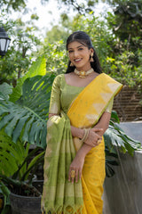 
A woman models a vibrant Golden Yellow Tussar Silk Saree with small woven motifs. The pallu features wide bands of contrasting olive green and horizontal gold Zari stripes. She wears a light green blouse and heavy gold Kundan jewelry, posing outdoors.