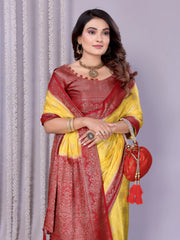 Woman wearing a yellow silk saree with a rich maroon embroidered border and matching blouse, styled with traditional jewelry. She poses gracefully in a contemporary studio featuring baskets filled with rose petals and minimal decor, set against a neutral backdrop.