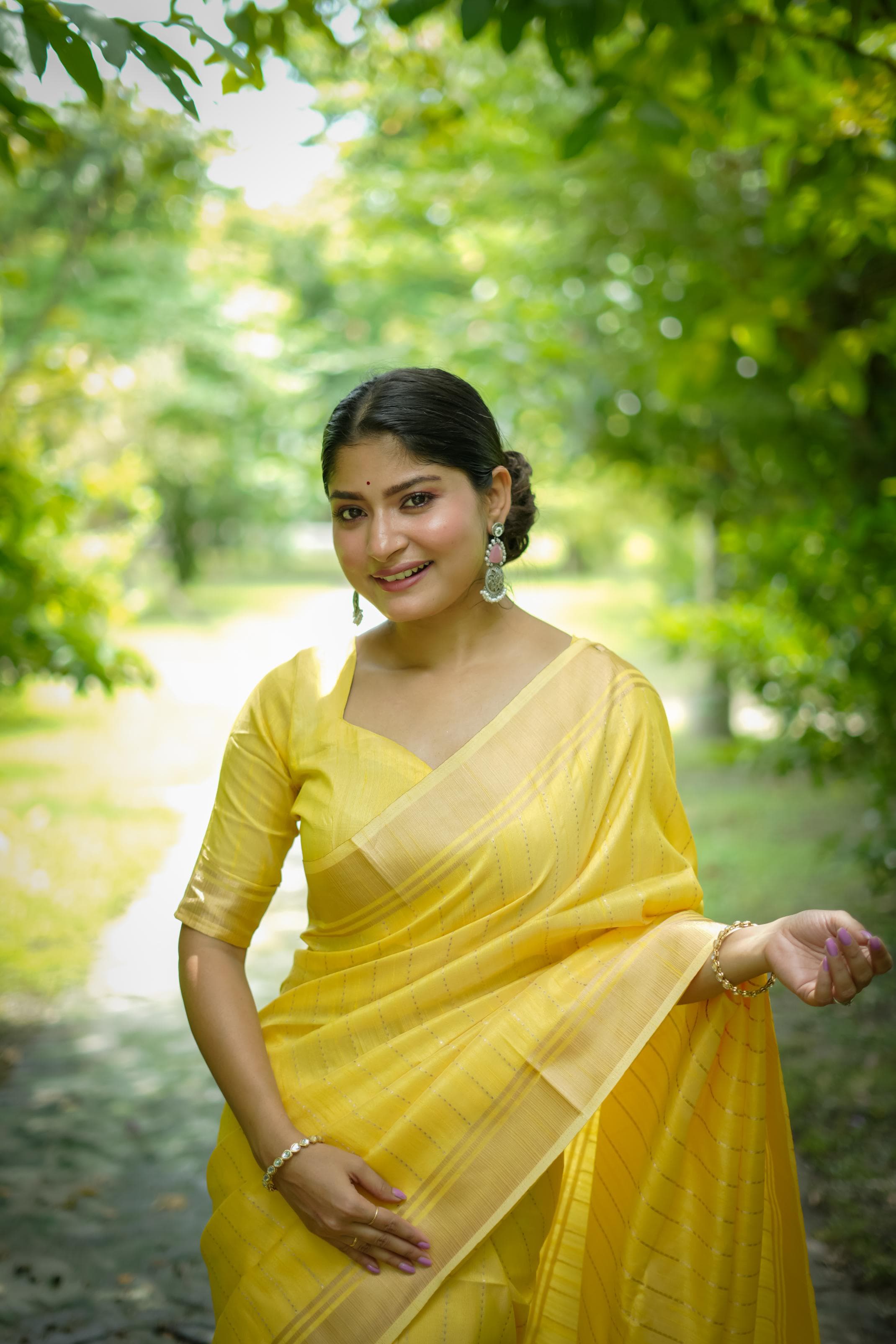 Woman in yellow zari work saree walking on a garden path surrounded by green trees and grass